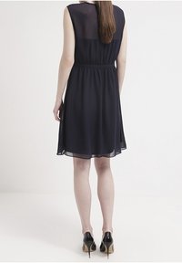 Abito senza maniche di un blu navy con una texture fluida e trasparente. Presenta una vita cinchata e una gonna svasata, abbinato a scarpe con tacco nero.