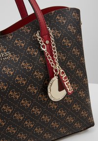 Tote bag met een gestructureerde zwarte synthetische buitenkant, voorzien van een bruin logo patroon, rode leren handvatten en een zilveren ketting met een bedel met merklogo.