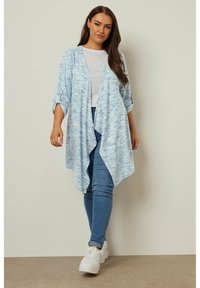 Ljusblå öppen cardigan med strukturerad stickad mönster, trekvartslånga ärmar och flödande silhuett, parat med åtsittande jeans och sneakers.