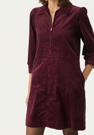 Femme portant une robe en velours côtelé bordeaux à manches longues et longueur genou, avec une fermeture éclair à l'avant et un col, les mains dans les poches latérales.