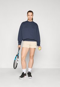 Marinefarbener oversized Sweatshirt, beigefarbene Sportshorts, hält einen Tennisschläger und einen grünen Ball, schwarze Sneakers, weiße Knöchelsocken. Minimalistisches Design.