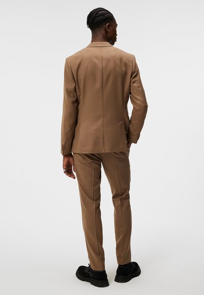 Brauner maßgeschneiderter Anzug mit einem taillierten Blazer und geraden Hosenbeinen, aus einem glatten Stoff gefertigt. Es werden schwarze Schuhe mit strukturierten Sohlen getragen.