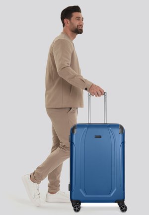 Blue hard-shell suitcase with a textured surface, retractable handle, and four wheels. Features rounded corner accents and a brand logo.