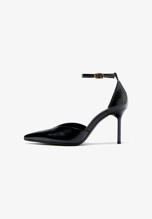 Schwarzer spitzer Lederpumps mit Knöchelriemen und goldener Schnalle, schlanker Stiletto-Absatz, Seitenansicht vor weißem Hintergrund.