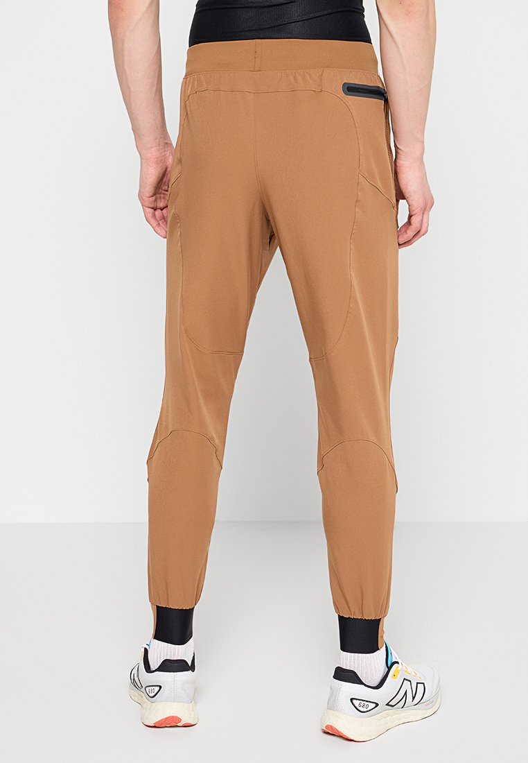 Pantalones deportivos marrones con piernas ajustadas, cinturilla elástica, bolsillos laterales y un bolsillo con cremallera. Se llevan con zapatillas gris y blanco.