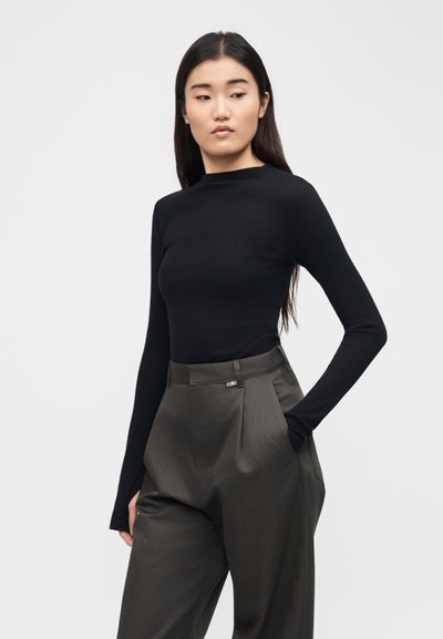 Γυναίκα με μακριά σκουρόχρωμα μαλλιά, φορώντας μαύρο μακρυμάνικο μπλουζάκι και ψηλόμεσο φαρδύ σκούρο γκρι παντελόνι, στέκεται με το ένα χέρι στην τσέπη.