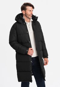 Sort pufferjakke med lang design, quiltet mønster, aftagelig hætte og trykknaplukning. Båret over en let sweater og jeans.