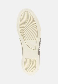 Podrážka topánky v off-white gumovom materiáli so vzorom v tvare zubov. Obsahuje sivý štítok s brandingom 'GUESS' na boku. Textúrovaná pre lepšiu priľnavosť.