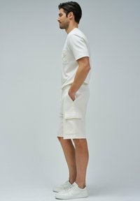 T-shirt en coton blanc avec logo embossé, associé à un short cargo blanc avec cordon de serrage, doté de poches latérales et de coutures plates. Baskets blanches.