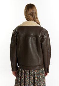 Giacca di pelle marrone con colletto in shearling crema, caratterizzata da una vestibilità rilassata, cuciture visibili e hardware in metallo. Indossata sopra un vestito floreale.