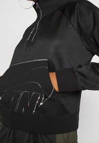 Svart zip-up sweatshirt med hög krage, med en reflekterande logotyp på framfickan och kontrasterande textilmaterial.