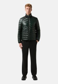 Tmavě zelená, polstrovaná bunda se zipem vpředu, vycházející z vysokého límce a geometrického šití. Nosit s černými kalhotami a botami.