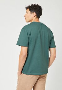 Jonge man met krullend donker haar, gekleed in een groene korte mouw shirt en beige corduroy broek, staande met de handen in de zakken en met de rug naar ons gekeerd.
