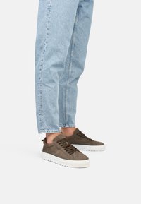 Zapatillas de ante marrón con parte superior texturizada, suela de goma blanca gruesa y cordones oscuros, llevadas con unos jeans ajustados de color azul claro.