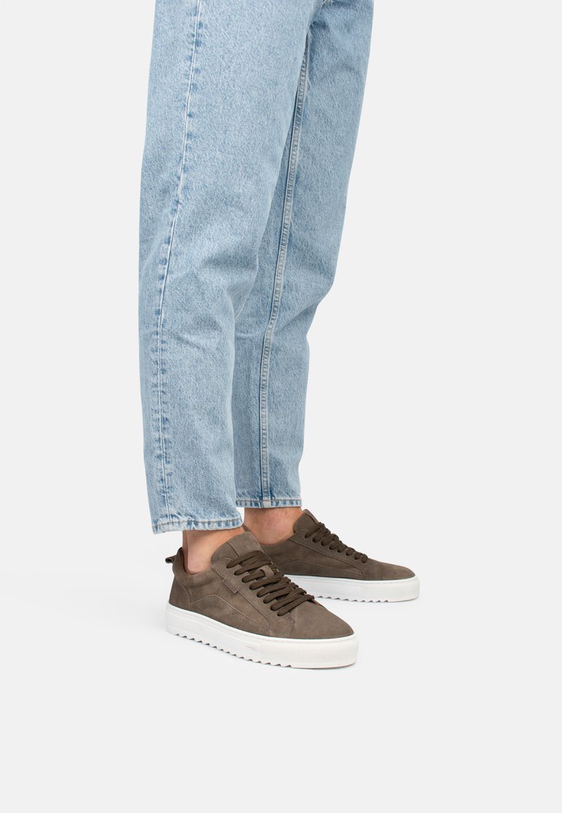 Zapatillas de ante marrón con parte superior texturizada, suela de goma blanca gruesa y cordones oscuros, llevadas con unos jeans ajustados de color azul claro.