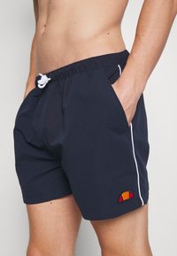 Torso masculino vestido con pantalones de baño azul marino con cordón blanco, bolsillos laterales, ribete blanco en los lados y un pequeño logotipo colorido cerca del dobladillo.