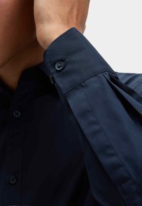 Navyblauwe shirt met een gladde textuur, voorzien van een knoopmanchet en geplooide accenten. Kraag en voor knopen zijn zichtbaar.