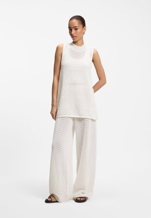 Femme portant une tunique en maille blanche sans manches et un pantalon à jambes larges, debout avec une main derrière le dos sur fond blanc.