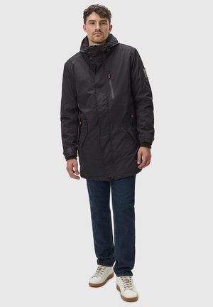 Zwarte waterdichte jas met een rits aan de voorkant, verstelbare capuchon, zijzakken en contrasterende oranje accenten. Draagt over een blauwe jeans en witte schoenen.