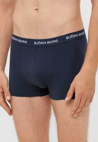 Temno modri boksarji iz mešanice mehkega bombaža, z belim logotipom na pasu, ki se kontrastno izstopa. Prilagojena zasnova z gladko teksturo.