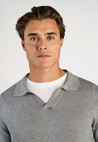 Grå stickad tröja med V-hals och krage, lagd över en vit T-shirt. Har en slät textur och figurformad passform.