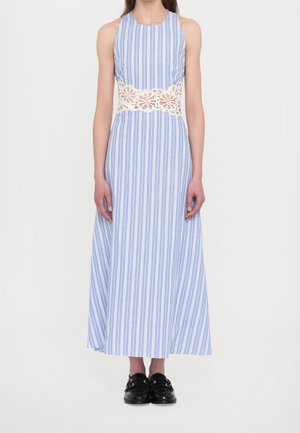Donna che indossa un vestito senza maniche a righe blu e bianco con un ritaglio in pizzo floreale bianco in vita e mocassini neri.