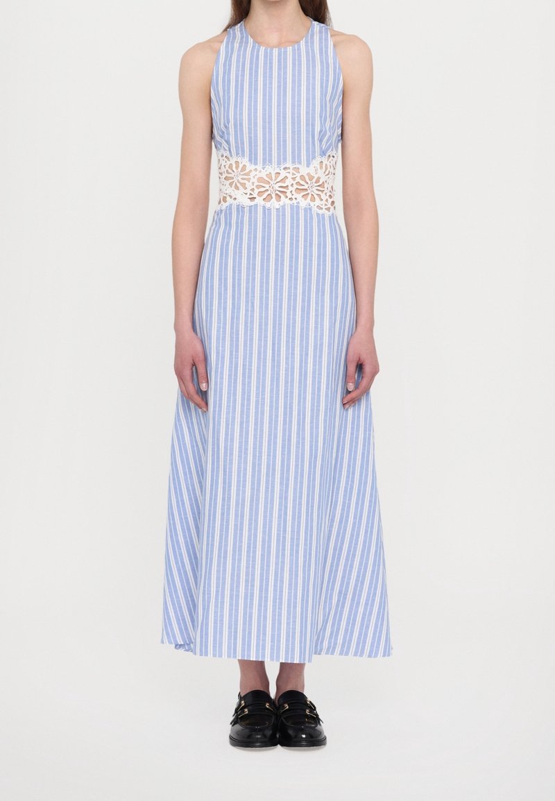 Femme portant une robe sans manches à rayures bleues et blanches avec une découpe en dentelle florale blanche à la taille et des mocassins noirs.