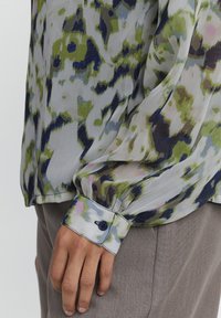 Blusa trasparente con un motivo astratto verde, blu e rosa. Maniche lunghe con polsini buttonati e una texture liscia.