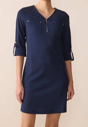 Mujer con un vestido azul marino hasta la rodilla, con mangas tres cuartos, cremallera frontal y dos bolsillos en el pecho con detalles de botones.