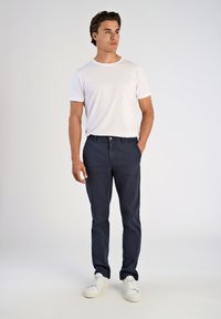 Ung man står med händerna i fickorna, klädd i en vit t-shirt med kort ärm, marinblå byxor och vita sneakers mot en neutral bakgrund.