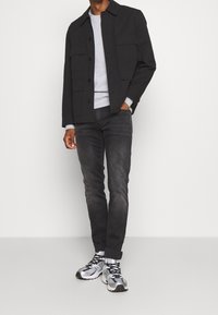 Chaqueta negra de botones con bolsillos en el pecho, suéter gris de cuello redondo, jeans oscuros y zapatillas negro plateadas con cordones y suelas de goma.