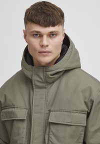 Oliven grønn jakke med hette, med en glatt tekstur, frontlukking med glidelås, og to store klaftlommer.