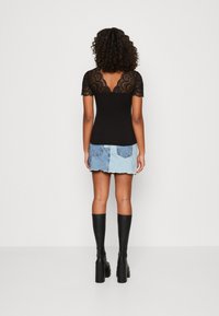 Schwarze Spitze-Bluse mit kurzen Ärmeln, die einen gewellten Ausschnitt und eine taillierte Passform hat. Getragen mit einem hellblauen Jeans-Minirock.