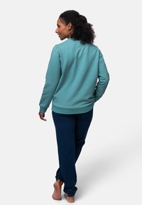 Felpa turchese con colletto rotondo e polsini a coste, abbinata a pantaloni jogger blu navy. Il materiale sembra morbido e informale.