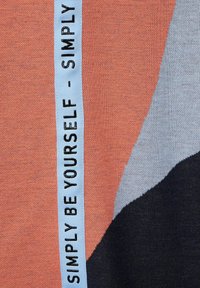 Färgblockerat tyg med orange, grå och svarta sektioner; ett blått band med texten "VARA DIG SJÄLV" tryckt i svart är tydligt framhävt.