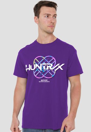 Junger Mann trägt ein violettes T-Shirt mit einem bunten geometrischen Logo und den Worten "HUNTRX" und "K-POP DEMON-HUNTERS" darauf.
