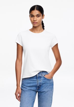 Femme aux cheveux bruns attachés en queue de cheval, portant un t-shirt blanc uni à manches courtes et un jean bleu, debout avec une main dans la poche.