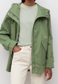 Osoba ubrana w jasnozieloną kurtkę na jasnoniebieskiej bluzie oraz ubrana w off-white spodnie, mająca jedną rękę w kieszeni.