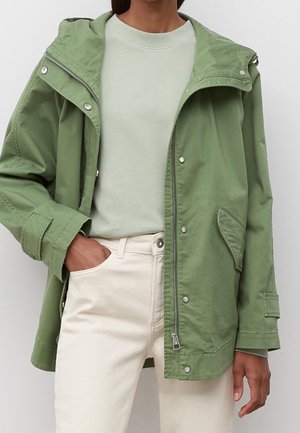 Persoon in een lichtgroene jas over een bleekgroene sweatvest en crèmekleurige broek, met één hand in de zak.