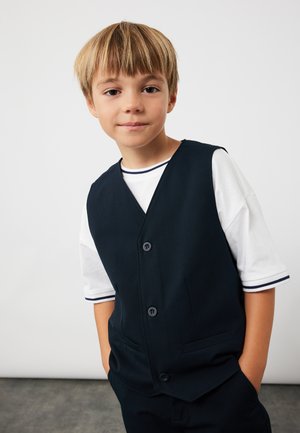 Marineblaues Westchen mit fünf Knöpfen, taillierte Passform, Taschen an der Vorderseite, getragen über einem weißen T-Shirt mit marinen gestreiften Säumen. Schlichter Hintergrund.