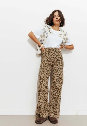Femme posant portant un t-shirt blanc, un gilet duveteux fleuri, un pantalon taille haute à imprimé léopard et des chaussures marron cloutées devant un mur blanc.