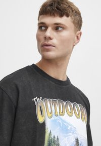 Svart oversized t-skjorte med et fjellmotiv med trær. Teksten "The OUTDOOR" er trykt med fet, stilisert skrift.