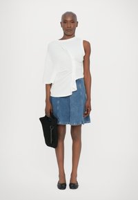 Biely top s krátkymi rukávmi, ktorý je drapovaný, v kombinácii s modrou džínovou sukňou. Modelka drží čiernu tkanú kabelku a má na sebe čierne ploché topánky.