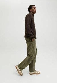 Maglione a coste marrone scuro con colletto alto, abbinato a pantaloni a gamba larga verde oliva e sneakers color crema con accenti marroni.