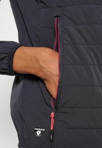 Veste matelassée isolante de couleur marine, avec un design matelassé, dotée d'une poche zippée avec des accents roses et un logo Primaloft. Tissu doux et texturé.