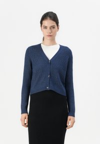 Navy knitted cardigan with a v-neck, ribbed texture, long sleeves, and heart-shaped buttons, layered over a white top and black skirt.