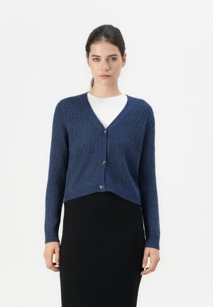 Navy gebreide cardigan met een v-hals, geribbelde textuur, lange mouwen en hartvormige knopen, gedragen over een witte top en een zwarte rok.