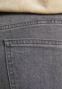 Gros plan d'une poche arrière sur un jean en denim gris avec des coutures visibles et un tissu beige au-dessus de la ceinture.