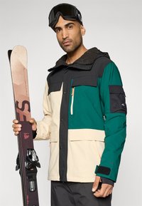 Casaco de esqui em verde, preto e bege, com múltiplos bolsos, capuz e punhos ajustáveis. Segura esquis e usa óculos de esqui pretos.