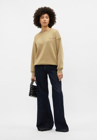 Khaki mikina s volným střihem a ribovanými manžetami, spárovaná s tmavě modrými rozšířenými džíny. Model drží malou černou tašku se stříbrnými akcenty.